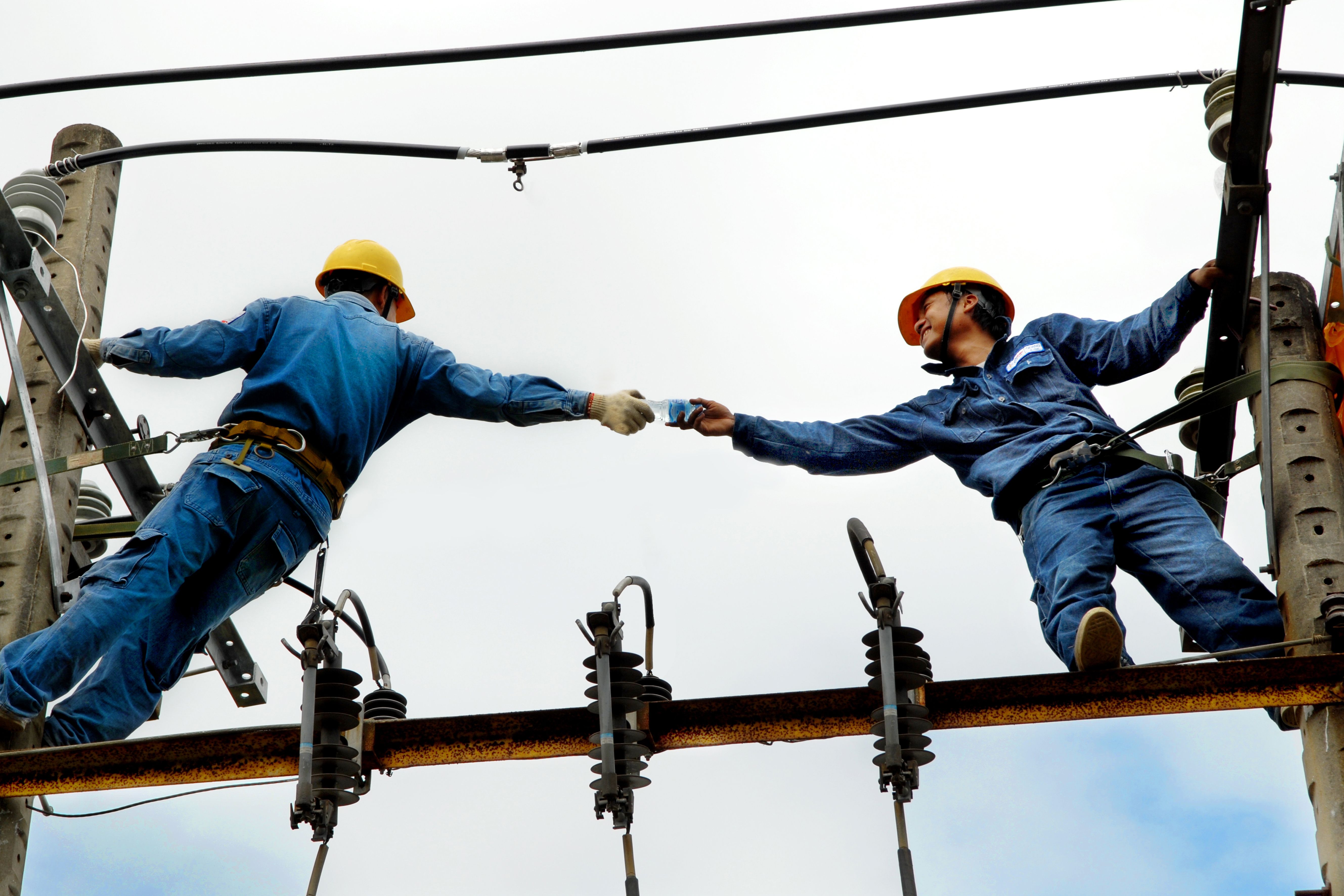 Two field technicians in harnesses working from an overhead line structure, passing equipment between them