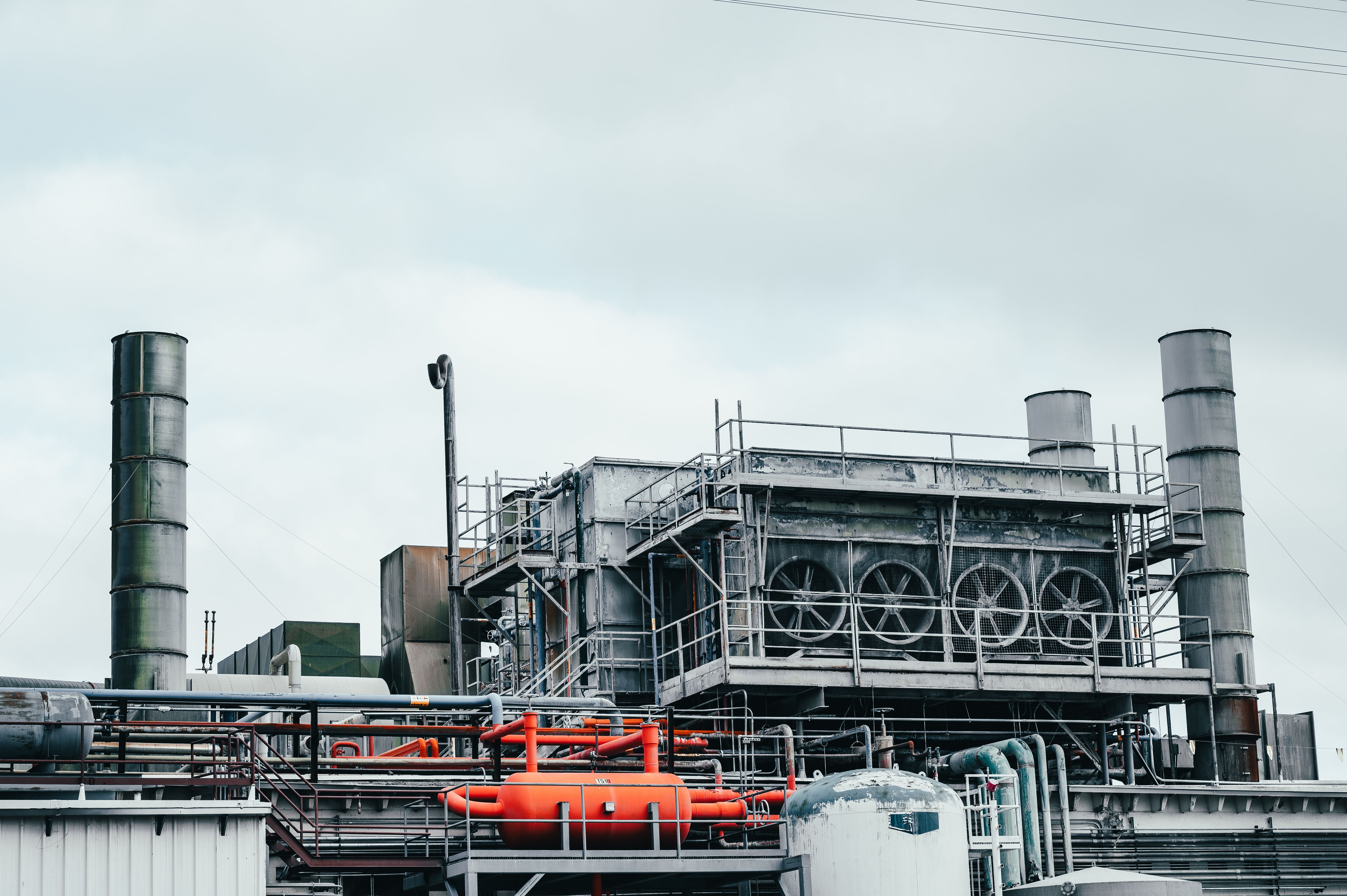 Industrial cooling plant with exhaust stacks, fan units and orange-painted piping under an overcast sky